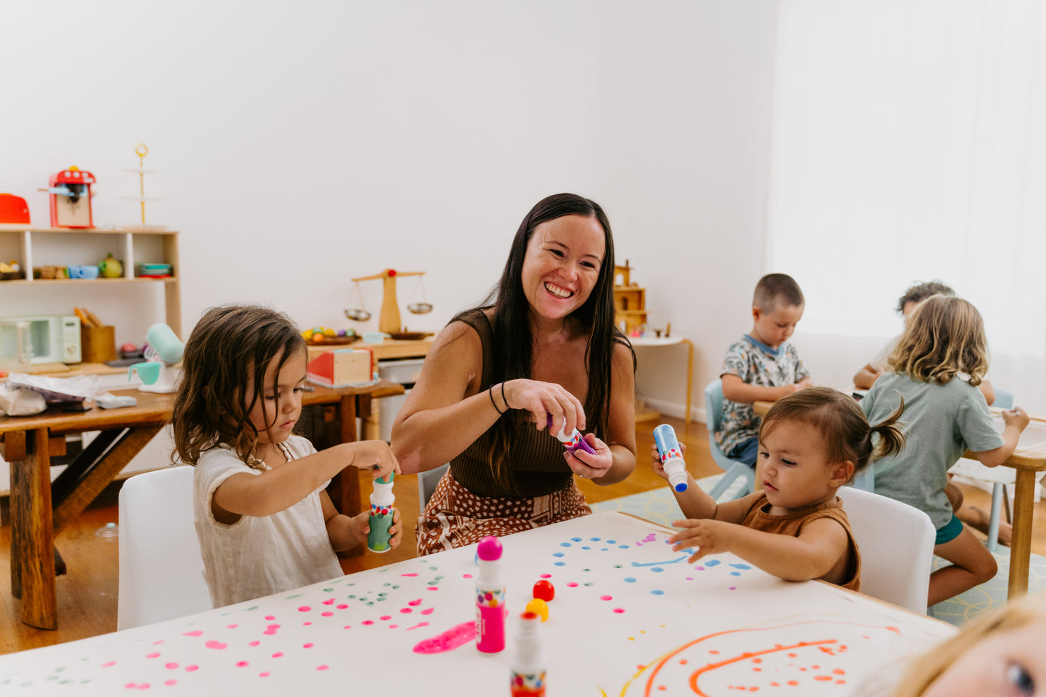 Teacher and young children engaged in a colourful painting activity in a bright classroom.