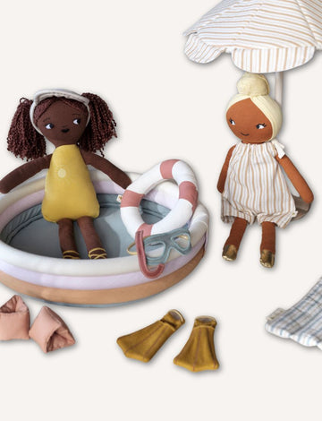 Two Fabelab Doll, Outfit & Accessories Bundles are near a small inflatable pool. One doll wears a yellow swimsuit, sitting inside with removable goggles and lifebuoy. The other stands nearby in a striped dress. Nearby are swim fins, arm floats, and a striped umbrella.