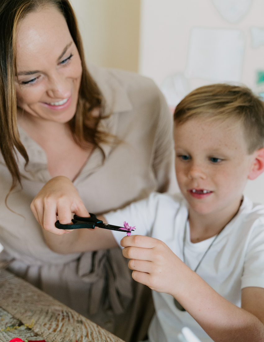 Woman assisting a young boy with fine motor skills using scissors and a flower.