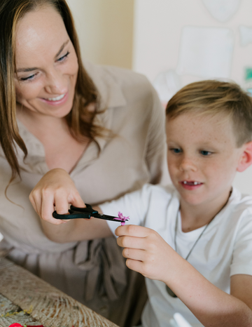 An adult and a child are engaged in the Fine Motor Skills Masterclass by The Play Way. The adult is holding scissors and guiding the child's hands as they cut a small, purple flower. Both appear to be focused on the task. They are indoors, and various art supplies from The Play Way are visible on the table.