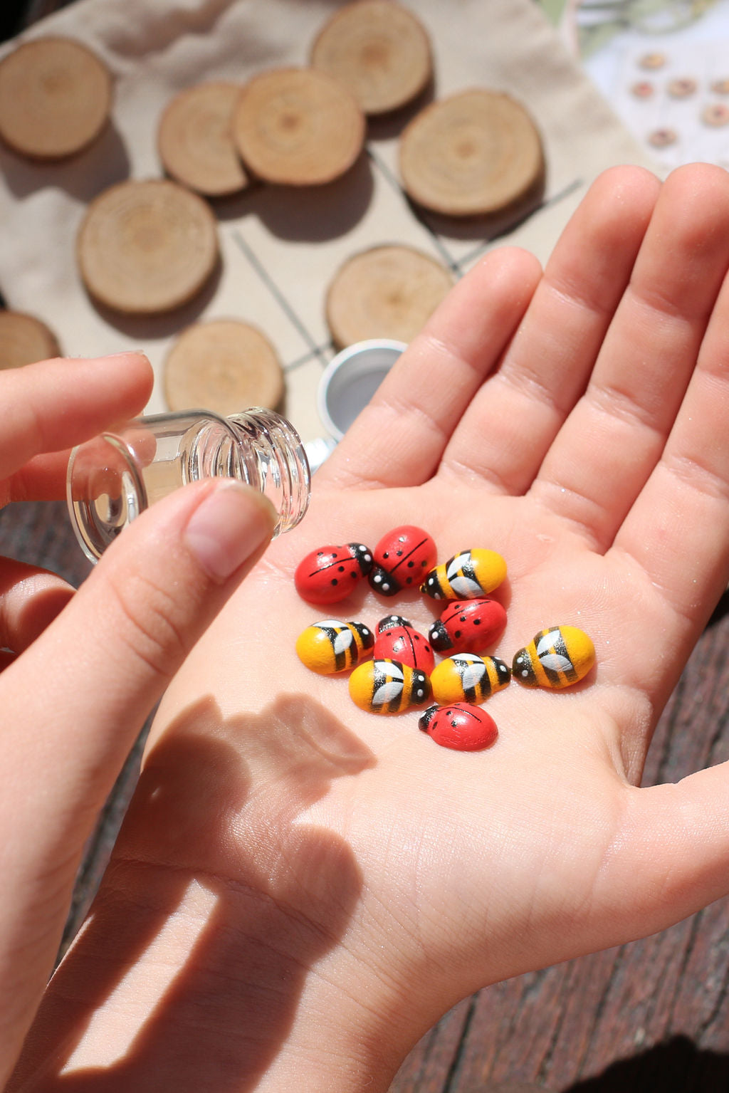 A hand displays small painted stones from the Poppy & Daisy DIY Tic Tac Toe set, featuring designs of ladybugs and bees, while another hand pours them from a small glass bottle. Circular wooden slices serve as a charming backdrop.