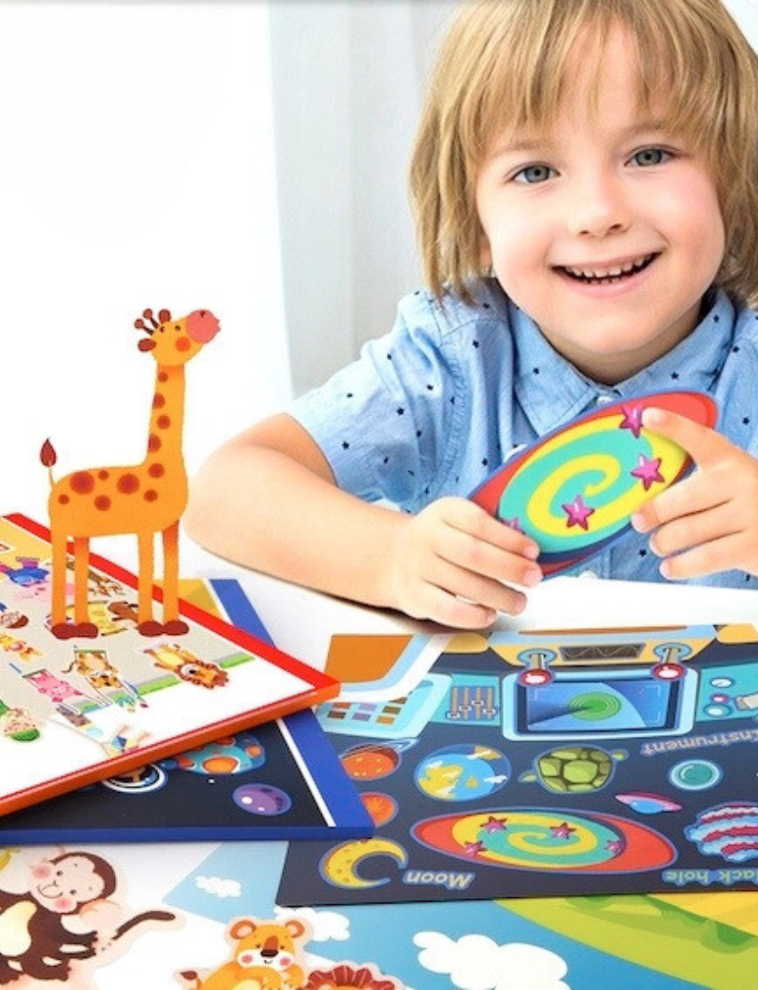 A child with light hair sits at a table, smiling and holding a colorful object. The table is decorated with bright, playful stickers from the Reusable Sticker Pad - City Park by Jar Melo, including a giraffe cut-out and various animal and space-themed designs.