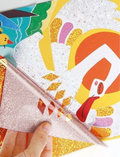 A close-up of a hand peeling back a sparkly, translucent layer from the vibrant Flash Stickers - The Ugly Duckling by Jar Melo. The artwork features abstract shapes and characters in dominant shades of orange, red, and yellow, with a shimmering texture overlay.