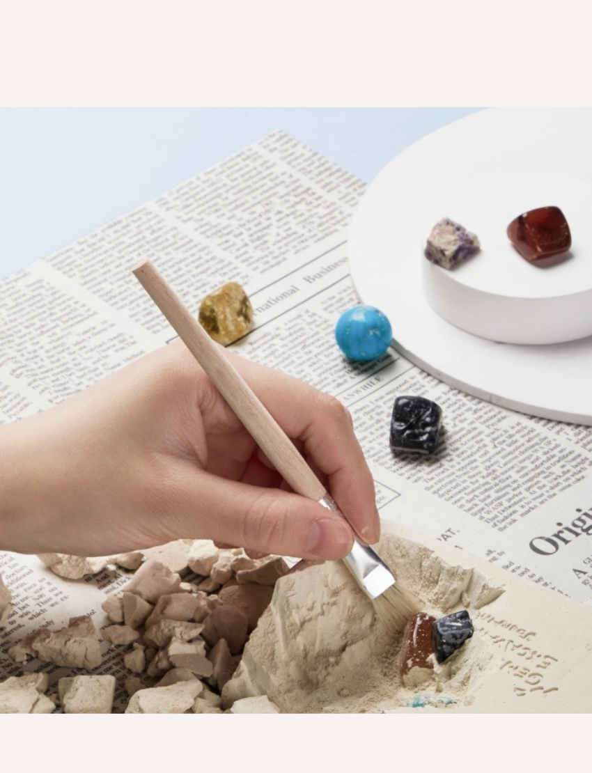 The image depicts a "Gemstone Excavation" STEM Archaeologist Kit by Jar Melo, intended for children aged 6 and older. The box showcases vibrant illustrations of a character unearthing various gemstones. It emphasizes "art, play, think" on the packaging and includes a safety warning.