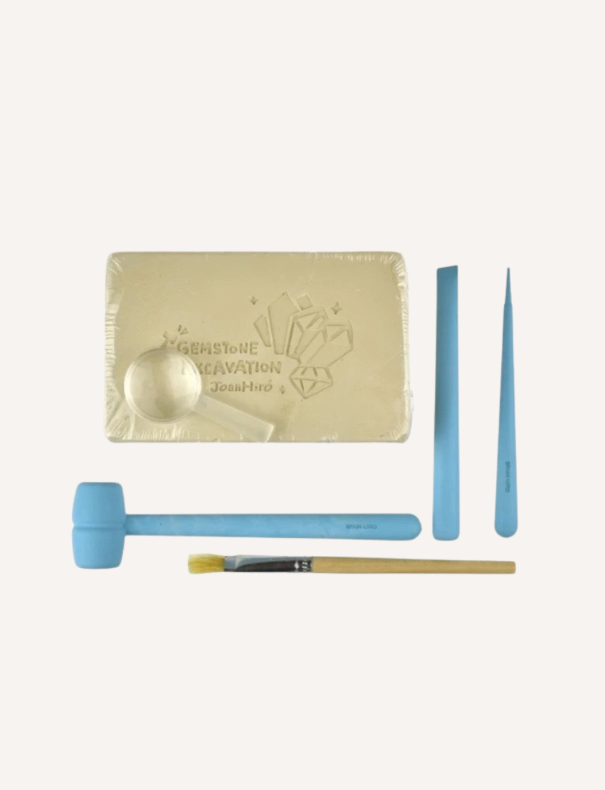 The image depicts a "Gemstone Excavation" STEM Archaeologist Kit by Jar Melo, intended for children aged 6 and older. The box showcases vibrant illustrations of a character unearthing various gemstones. It emphasizes "art, play, think" on the packaging and includes a safety warning.