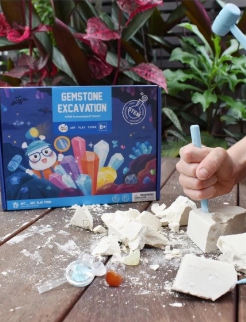 The image depicts a "Gemstone Excavation" STEM Archaeologist Kit by Jar Melo, intended for children aged 6 and older. The box showcases vibrant illustrations of a character unearthing various gemstones. It emphasizes "art, play, think" on the packaging and includes a safety warning.