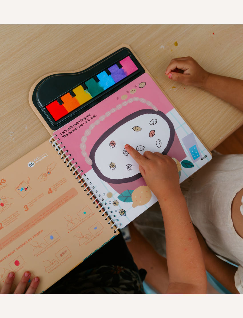 Two children explore a page from the Finger Painting - A Lively Forest book by Jar Melo, showcasing a vibrant xylophone. One child points at small images of acorns and leaves featured on the page. The book offers instructions and bold, colorful shapes to aid learning.