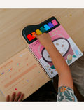 Two children explore a page from the Finger Painting - A Lively Forest book by Jar Melo, showcasing a vibrant xylophone. One child points at small images of acorns and leaves featured on the page. The book offers instructions and bold, colorful shapes to aid learning.