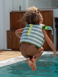 A child in a pool wearing the Sunnylife Kids Swim Vest Into The Wild in khaki, reaching out to a crocodile-shaped float with big eyes.