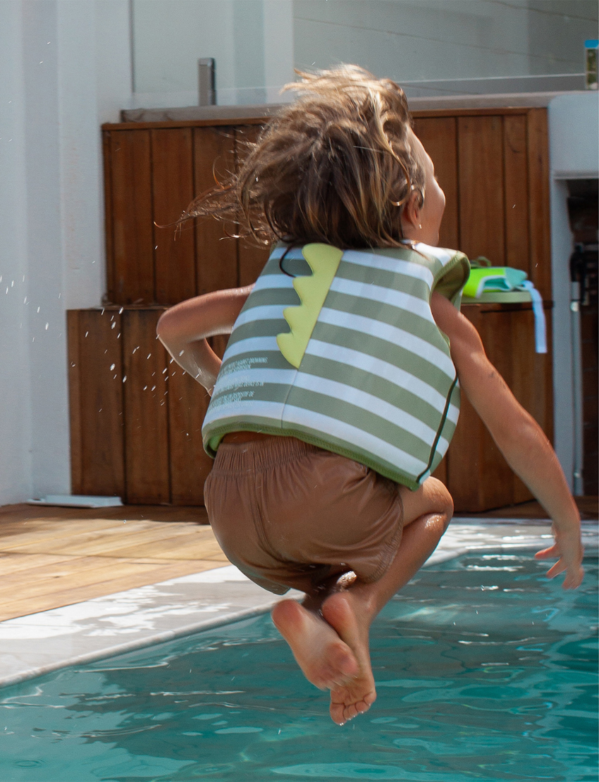 A child in a pool wearing the Sunnylife Kids Swim Vest Into The Wild in khaki, reaching out to a crocodile-shaped float with big eyes.