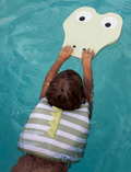 A child in a pool wearing the Sunnylife Kids Swim Vest Into The Wild in khaki, reaching out to a crocodile-shaped float with big eyes.