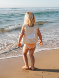 A young child with blonde hair sits on a sandy beach, wearing a Sunnylife Kids Swim Vest Princess Swan in Yellow. They are playing with the sand, and the ocean is visible in the background under a clear sky.