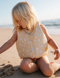 A young child with blonde hair sits on a sandy beach, wearing a Sunnylife Kids Swim Vest Princess Swan in Yellow. They are playing with the sand, and the ocean is visible in the background under a clear sky.