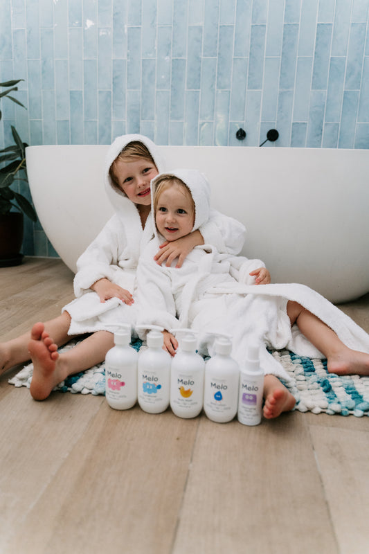 A teal box adorned with colorful shapes opened to reveal the Melo Self-Care Starter Kit by The Play Way, featuring body lotion, body wash, shampoo, conditioner, and detangling spray. Each item is packaged in a white bottle with various illustrations and labeled "Melo.