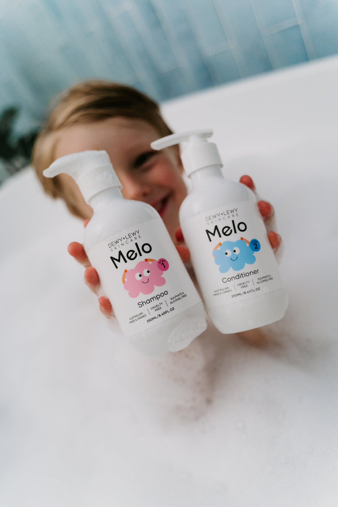 Two white pump bottles of Melo Shampoo by Melo are shown. The front of one bottle features a smiling pink character and the text “Melo Shampoo,” while the back of the other bottle displays ingredients and usage instructions in black text. This gentle, cruelty-free shampoo is perfect for sensitive scalps.