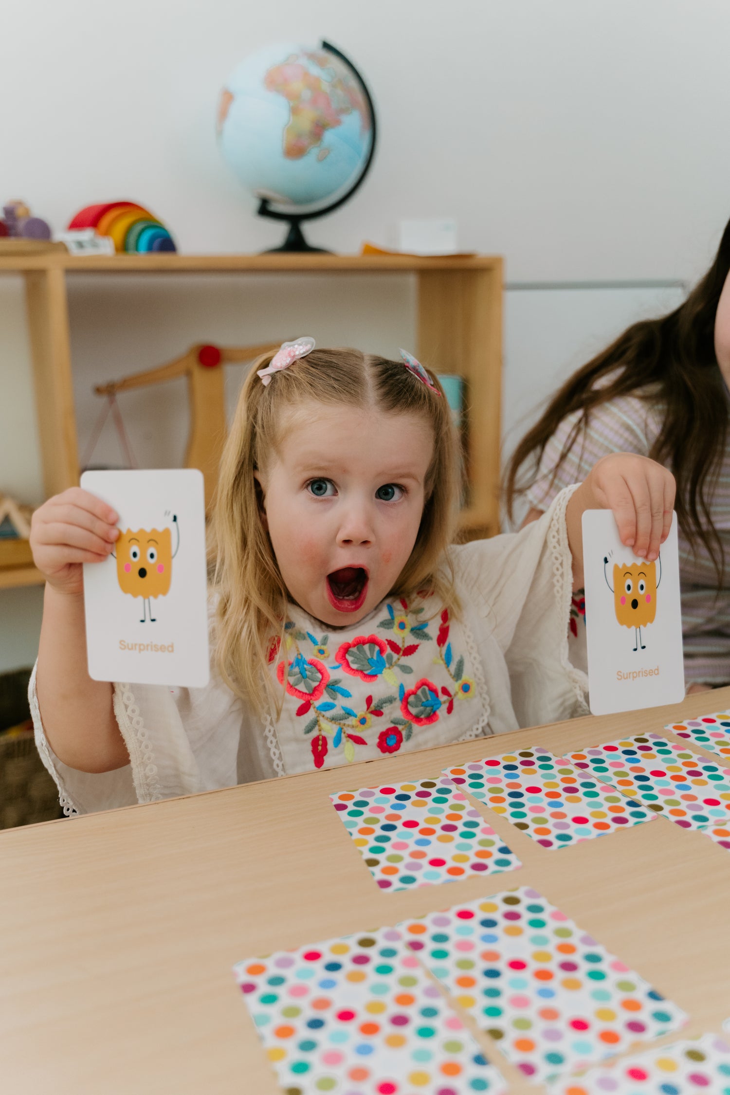 A surprised young girl holds up cards showing the emotion 'Surprised', engaging in an emotional literacy activity.