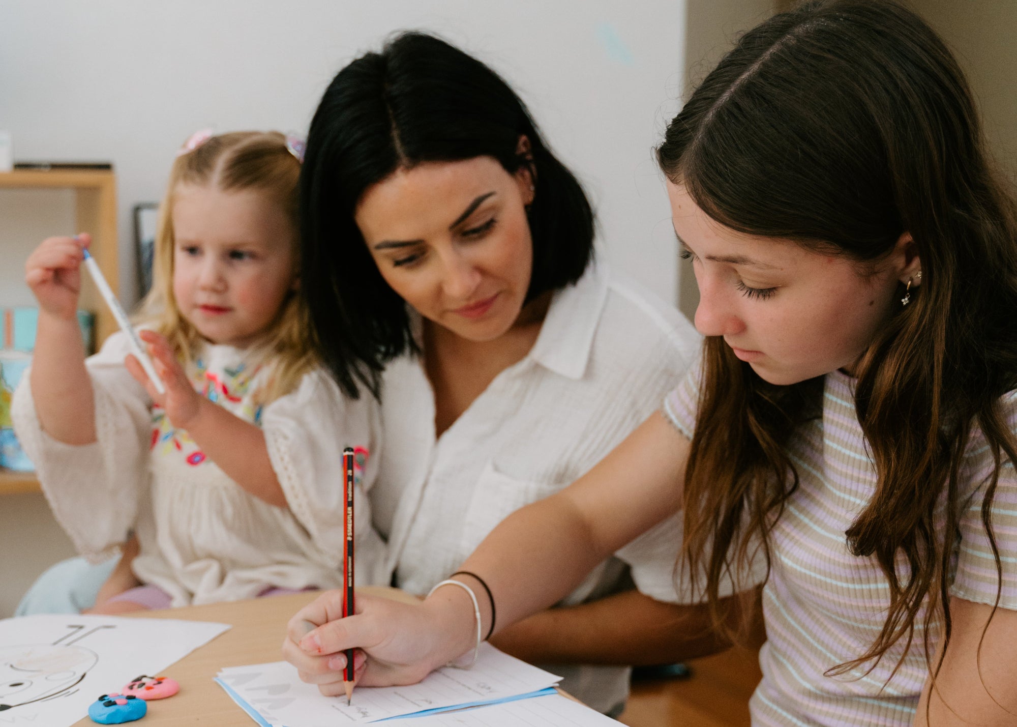 A woman and two girls sit at a light wood table in a home setting, working on papers. The woman in the center holds a red pen while writing, with the girls looking down at the sheets.