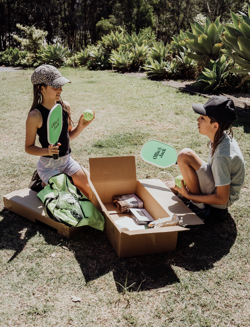 Two kids are smiling and posing outdoors with their arms around each other while holding paddles and balls from the Ollie & Jack LED Pickleball Net Set. They are dressed in casual summer clothing and caps, with greenery in the background beneath a sunny sky.