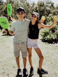 Two kids are smiling and posing outdoors with their arms around each other while holding paddles and balls from the Ollie & Jack LED Pickleball Net Set. They are dressed in casual summer clothing and caps, with greenery in the background beneath a sunny sky.