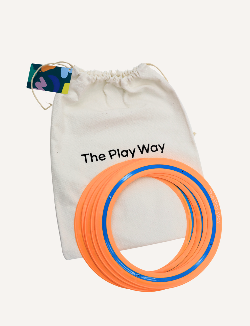 A freckled child with curly hair excitedly holds up an orange and blue Frisbee - Pack of 5 from The Play Way, in front of their face. Outdoors in a denim jacket, they smile amid the outdoor fun with blurred trees and grass adding a lively backdrop.