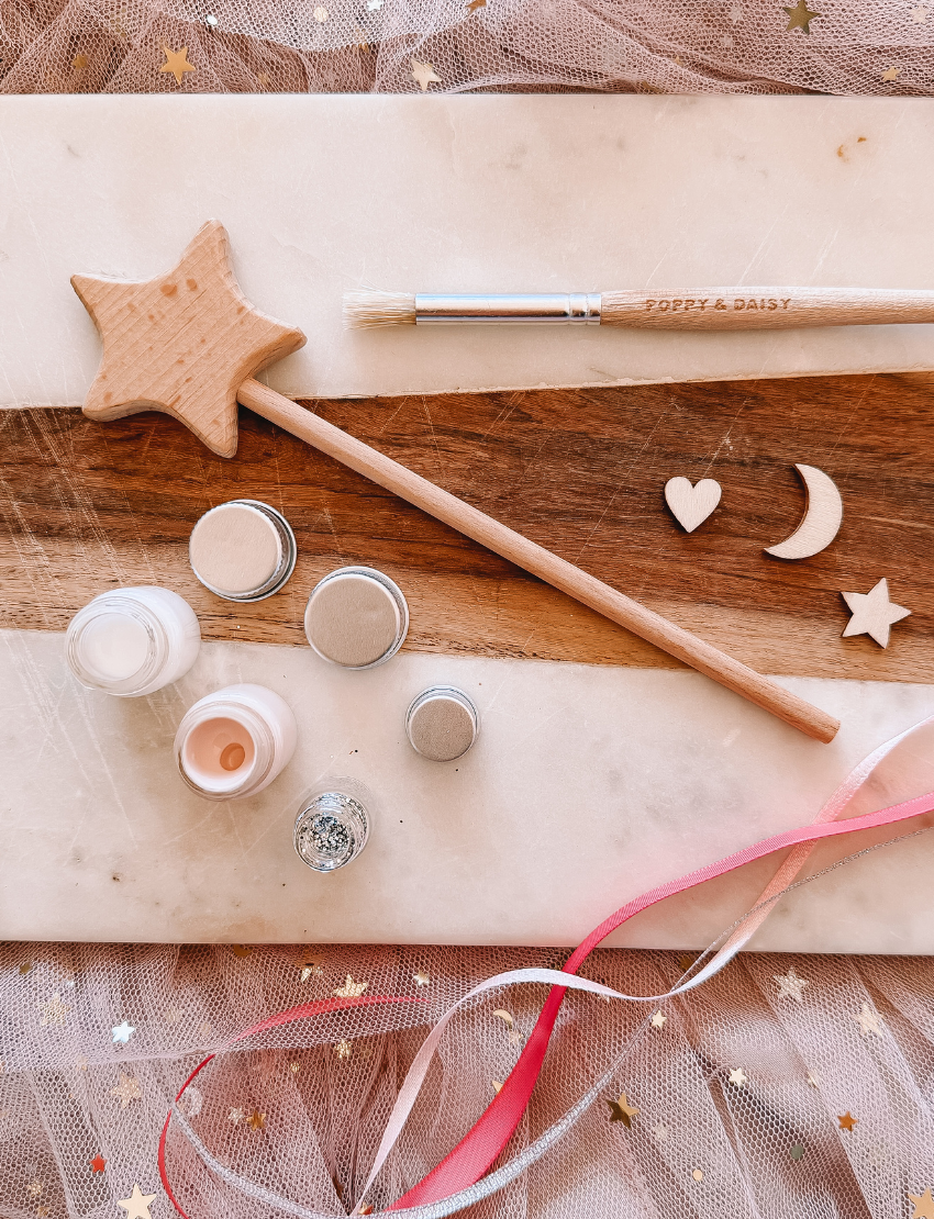 A flat lay image features a "DIY Fairy Wand" by Poppy & Daisy, accompanied by a brush with the brand name "Poppy & Daisy," small jars containing cream and glitter, and various wooden shapes like a heart, moon, and star. The arrangement is enhanced by pink and gold ribbons scattered across the textured surface.