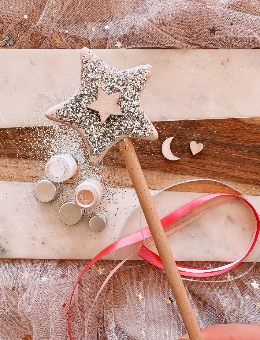 A flat lay image features a "DIY Fairy Wand" by Poppy & Daisy, accompanied by a brush with the brand name "Poppy & Daisy," small jars containing cream and glitter, and various wooden shapes like a heart, moon, and star. The arrangement is enhanced by pink and gold ribbons scattered across the textured surface.