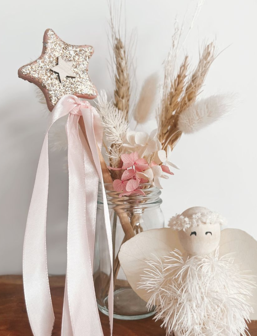 A flat lay image features a "DIY Fairy Wand" by Poppy & Daisy, accompanied by a brush with the brand name "Poppy & Daisy," small jars containing cream and glitter, and various wooden shapes like a heart, moon, and star. The arrangement is enhanced by pink and gold ribbons scattered across the textured surface.