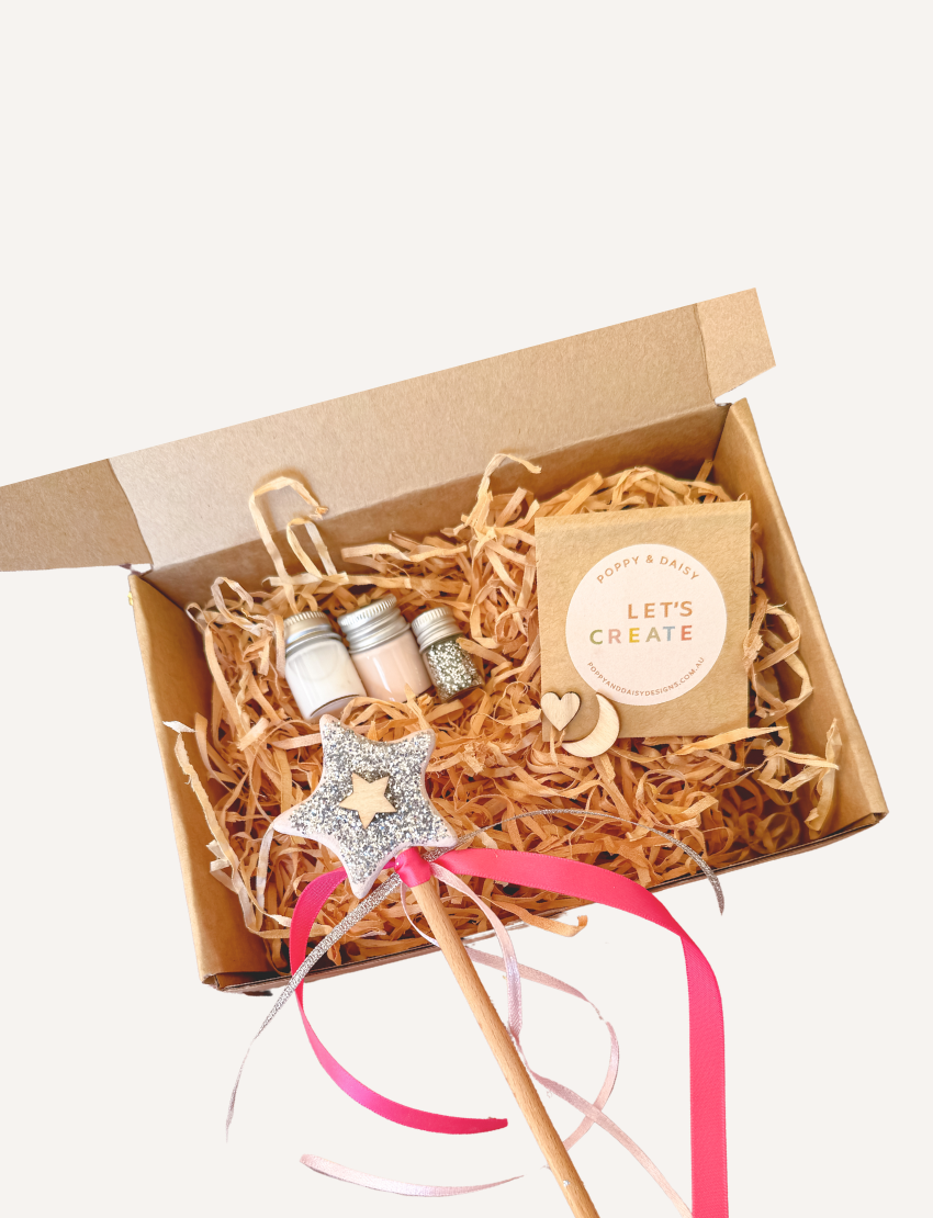 A flat lay image features a "DIY Fairy Wand" by Poppy & Daisy, accompanied by a brush with the brand name "Poppy & Daisy," small jars containing cream and glitter, and various wooden shapes like a heart, moon, and star. The arrangement is enhanced by pink and gold ribbons scattered across the textured surface.