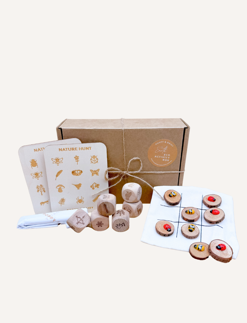 Two people are seated on a woven mat, enjoying their time with small wooden dice from the Poppy & Daisy Activity Box. The round wooden tray, which holds the dice adorned with various symbols, is complemented by dried leaves and pinecones artfully arranged beside it.