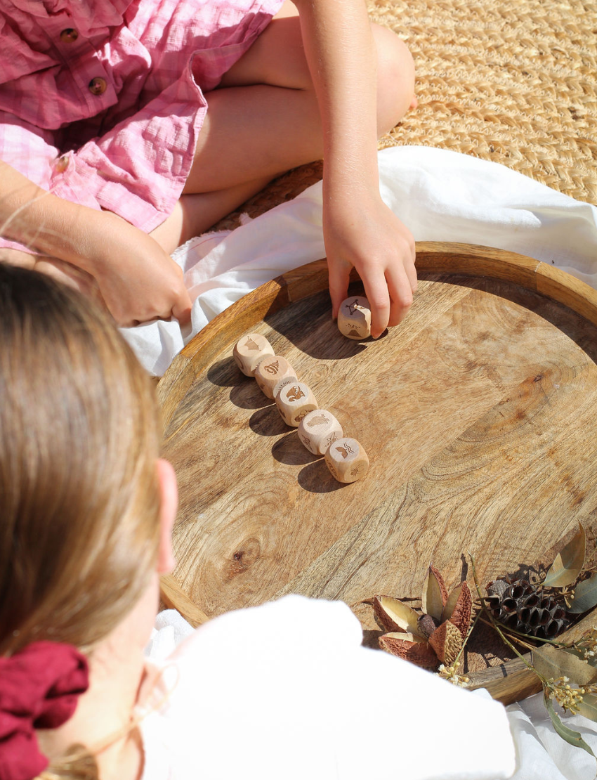 Two people are seated on a woven mat, enjoying their time with small wooden dice from the Poppy & Daisy Activity Box. The round wooden tray, which holds the dice adorned with various symbols, is complemented by dried leaves and pinecones artfully arranged beside it.