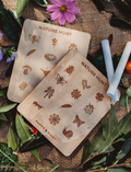 Two people are seated on a woven mat, enjoying their time with small wooden dice from the Poppy & Daisy Activity Box. The round wooden tray, which holds the dice adorned with various symbols, is complemented by dried leaves and pinecones artfully arranged beside it.