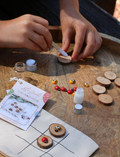 Two people are seated on a woven mat, enjoying their time with small wooden dice from the Poppy & Daisy Activity Box. The round wooden tray, which holds the dice adorned with various symbols, is complemented by dried leaves and pinecones artfully arranged beside it.