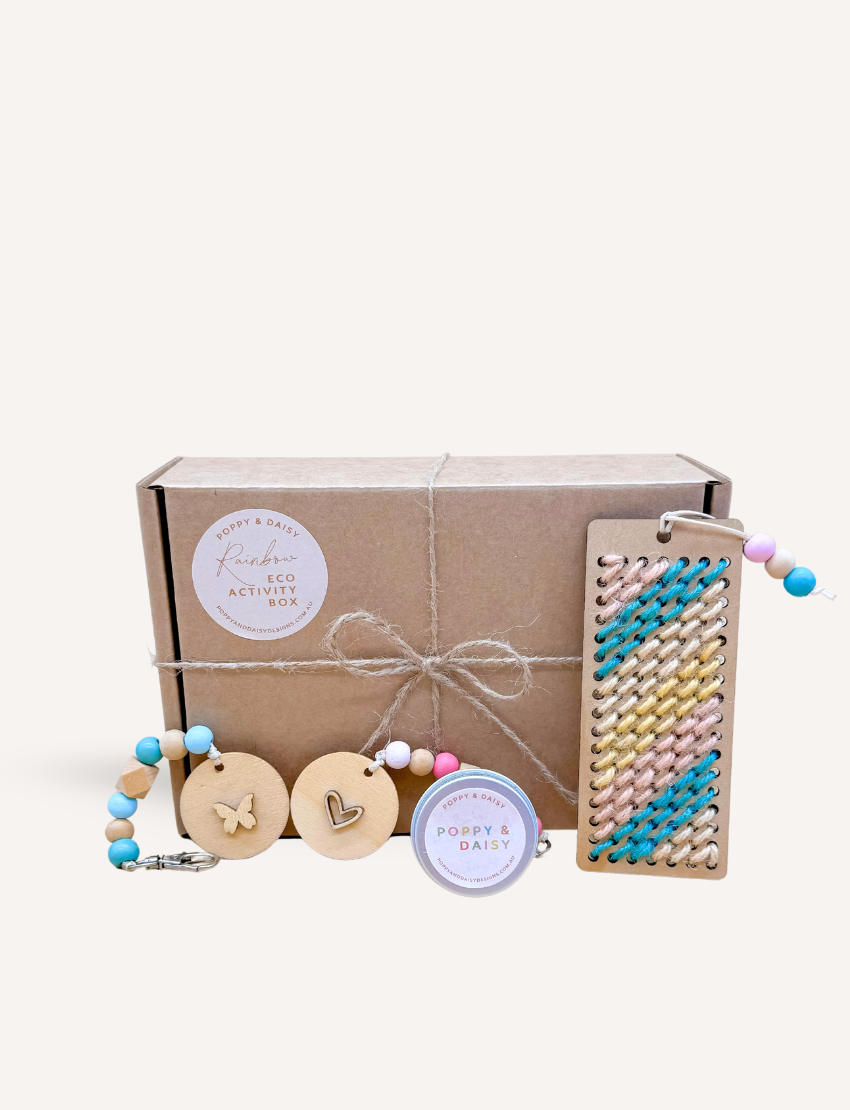 Two people are seated on a woven mat, enjoying their time with small wooden dice from the Poppy & Daisy Activity Box. The round wooden tray, which holds the dice adorned with various symbols, is complemented by dried leaves and pinecones artfully arranged beside it.