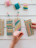 Two people are seated on a woven mat, enjoying their time with small wooden dice from the Poppy & Daisy Activity Box. The round wooden tray, which holds the dice adorned with various symbols, is complemented by dried leaves and pinecones artfully arranged beside it.