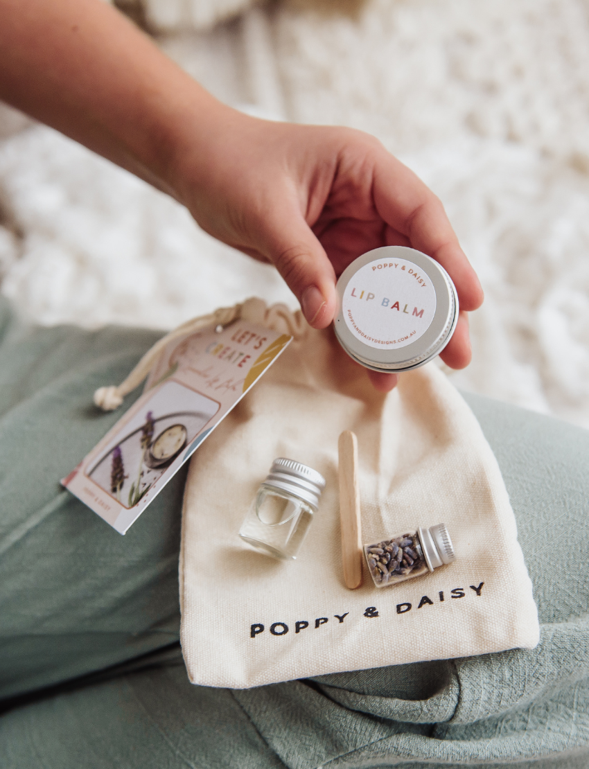 Two people are seated on a woven mat, enjoying their time with small wooden dice from the Poppy & Daisy Activity Box. The round wooden tray, which holds the dice adorned with various symbols, is complemented by dried leaves and pinecones artfully arranged beside it.