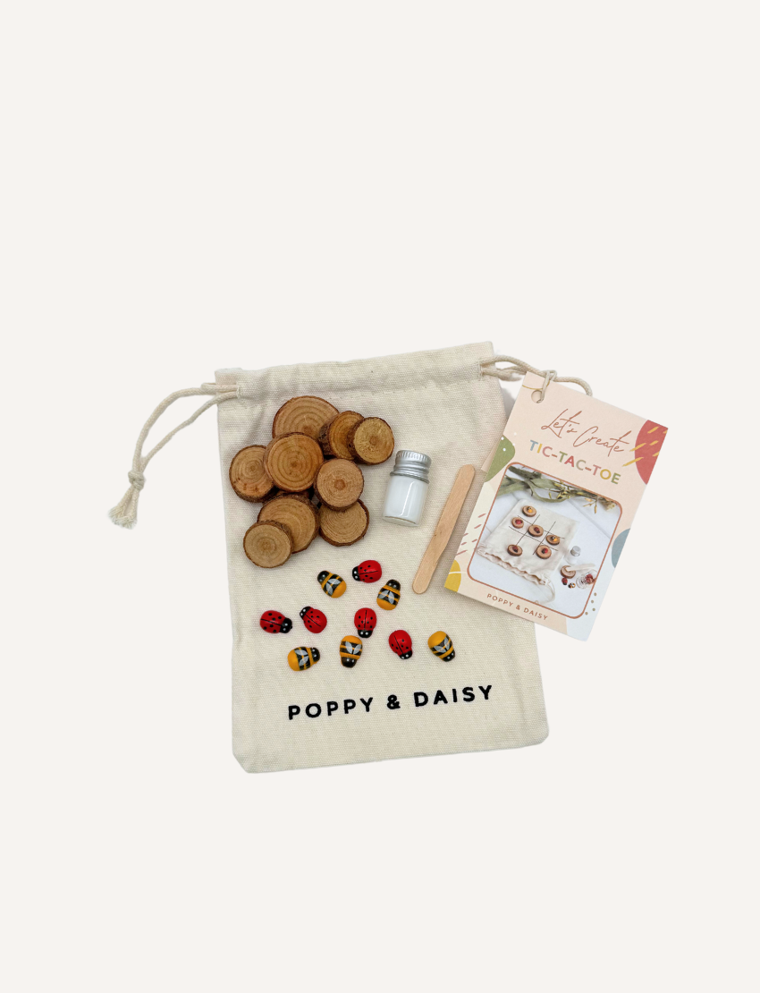 A hand displays small painted stones from the Poppy & Daisy DIY Tic Tac Toe set, featuring designs of ladybugs and bees, while another hand pours them from a small glass bottle. Circular wooden slices serve as a charming backdrop.