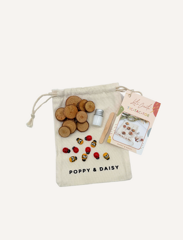 A hand displays small painted stones from the Poppy & Daisy DIY Tic Tac Toe set, featuring designs of ladybugs and bees, while another hand pours them from a small glass bottle. Circular wooden slices serve as a charming backdrop.