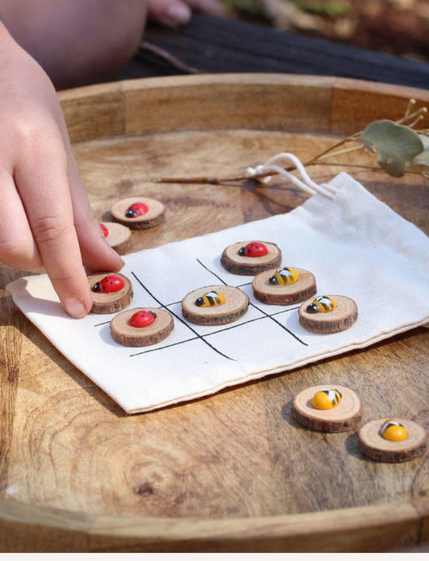 A hand displays small painted stones from the Poppy & Daisy DIY Tic Tac Toe set, featuring designs of ladybugs and bees, while another hand pours them from a small glass bottle. Circular wooden slices serve as a charming backdrop.