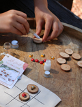 A hand displays small painted stones from the Poppy & Daisy DIY Tic Tac Toe set, featuring designs of ladybugs and bees, while another hand pours them from a small glass bottle. Circular wooden slices serve as a charming backdrop.