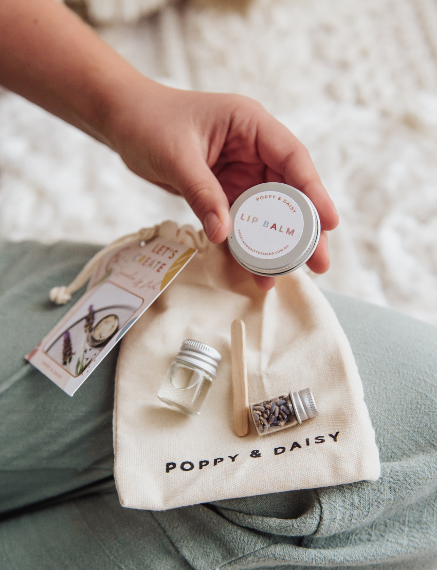 A hand holds a small tin of Lavender Lip Balm. Below it, a cloth pouch labeled "Poppy & Daisy" contains a glass vial, a small wooden stick, and a card with floral illustrations. The background reveals a knitted blanket.