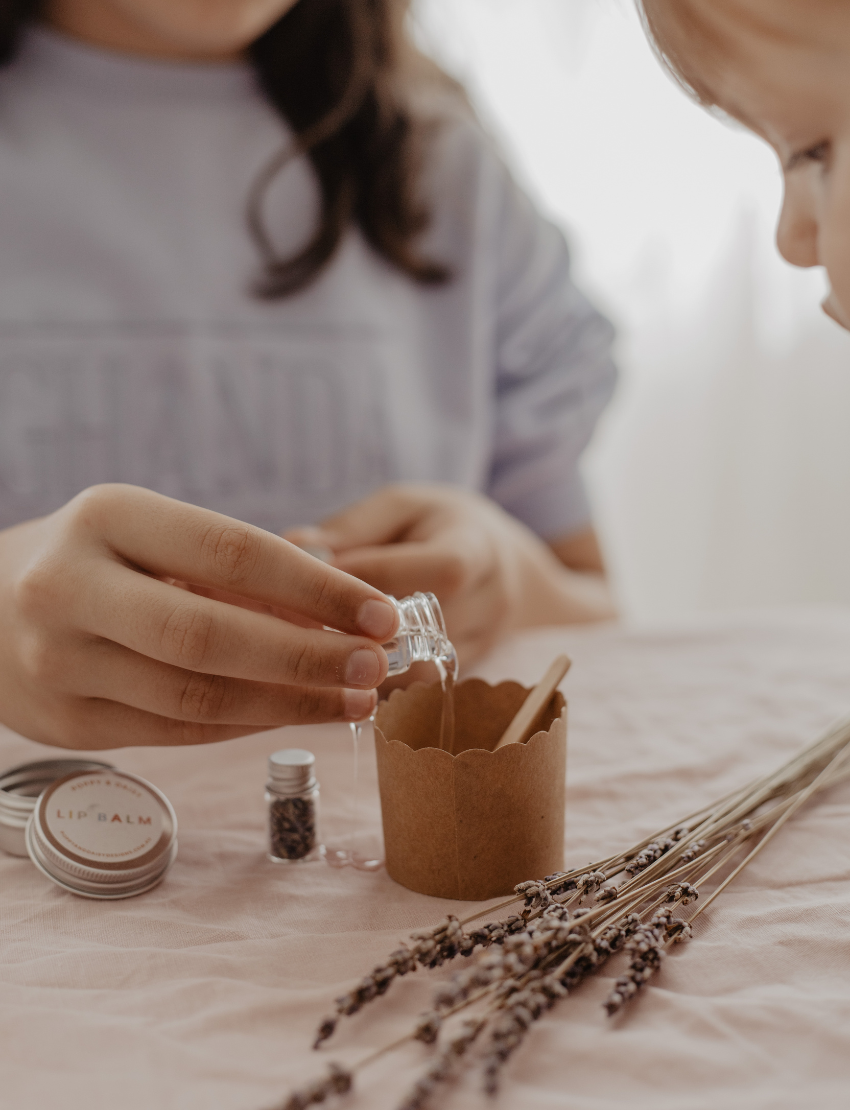 A hand holds a small tin of Lavender Lip Balm. Below it, a cloth pouch labeled "Poppy & Daisy" contains a glass vial, a small wooden stick, and a card with floral illustrations. The background reveals a knitted blanket.