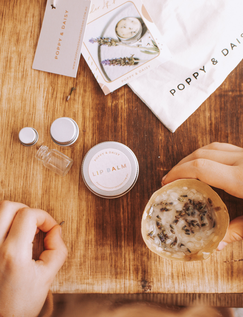 A hand holds a small tin of Lavender Lip Balm. Below it, a cloth pouch labeled "Poppy & Daisy" contains a glass vial, a small wooden stick, and a card with floral illustrations. The background reveals a knitted blanket.