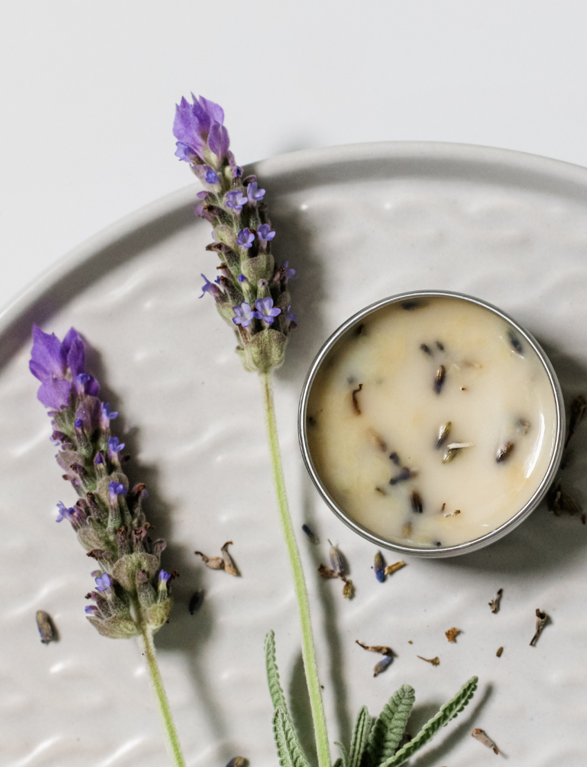 A hand holds a small tin of Lavender Lip Balm. Below it, a cloth pouch labeled "Poppy & Daisy" contains a glass vial, a small wooden stick, and a card with floral illustrations. The background reveals a knitted blanket.