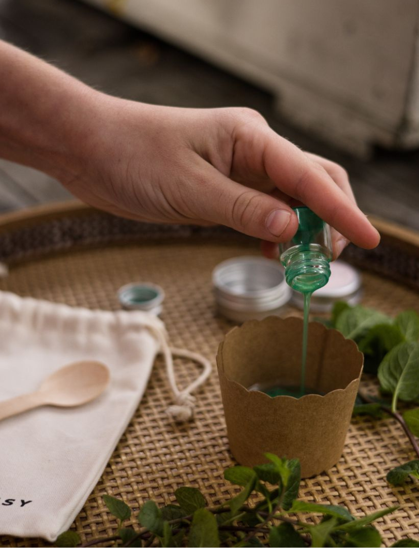 The Peppermint Lip Balm kit by Poppy & Daisy contains a cloth bag with the brand's label, a small jar of lip balm, a metal jar featuring a label, a wooden spoon, a scalloped paper cup, a small green bottle, and an instruction card complete with a photo.