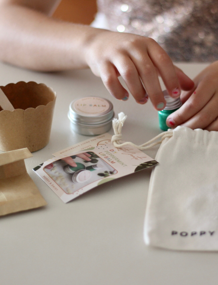 The Peppermint Lip Balm kit by Poppy & Daisy contains a cloth bag with the brand's label, a small jar of lip balm, a metal jar featuring a label, a wooden spoon, a scalloped paper cup, a small green bottle, and an instruction card complete with a photo.