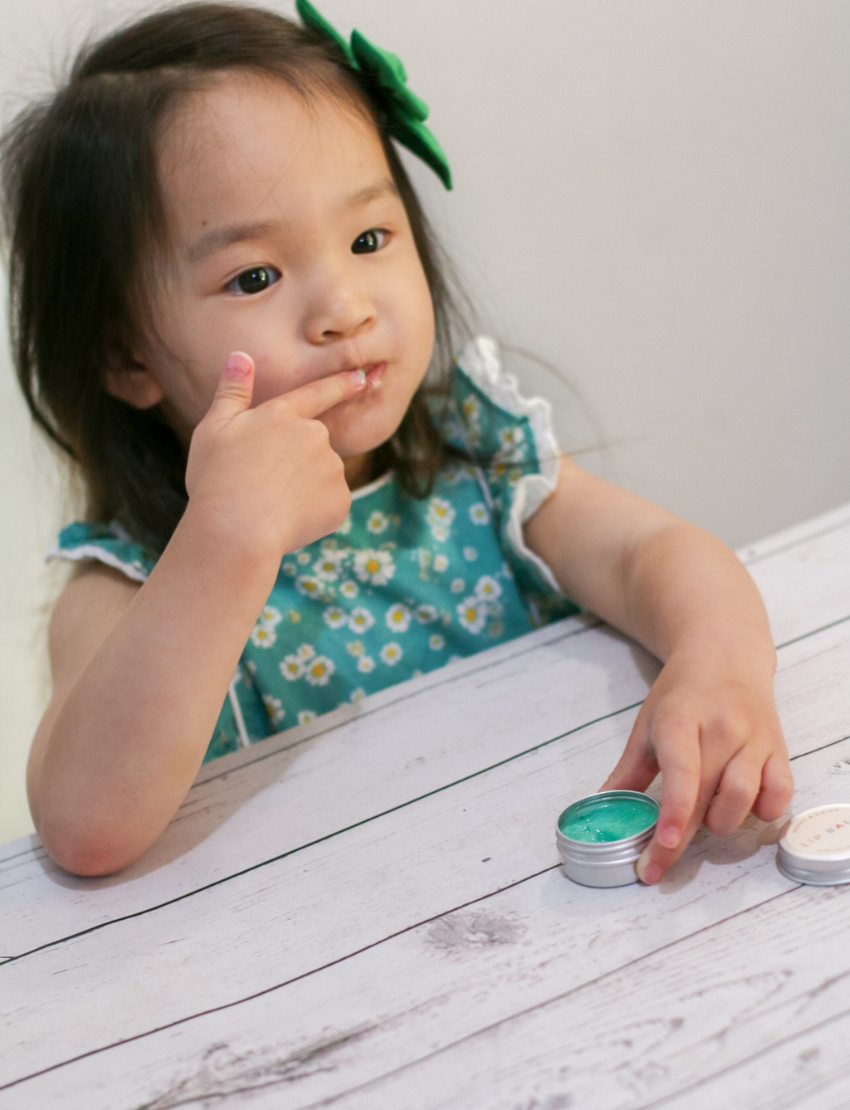 The Peppermint Lip Balm kit by Poppy & Daisy contains a cloth bag with the brand's label, a small jar of lip balm, a metal jar featuring a label, a wooden spoon, a scalloped paper cup, a small green bottle, and an instruction card complete with a photo.