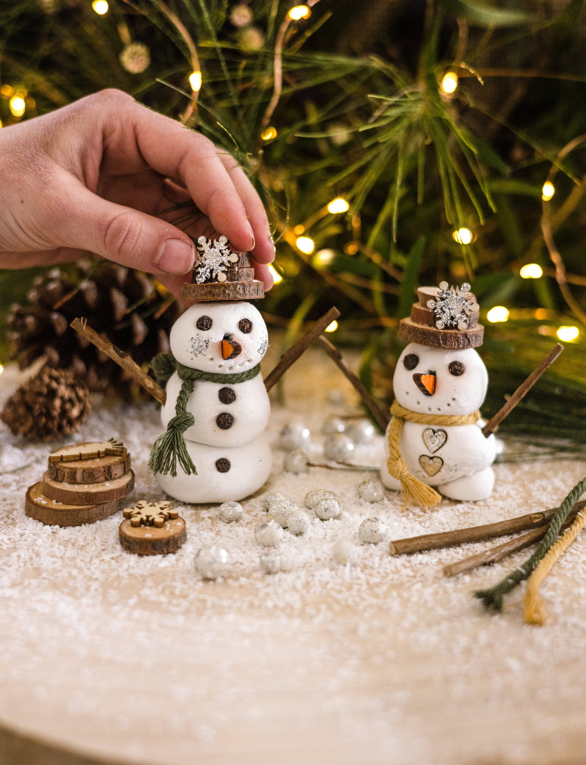 Introducing the Clay Snowmen kit by Poppy & Daisy, which comes in a "Let's Create" labeled cardboard box featuring an image of finished snowmen. The kit includes a tub of clay, sticks, yarn, wooden cutouts, and small containers filled with glitter and beads along with complete instructions.