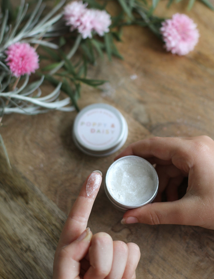 A person holds a small open tin of Natural Body Glitter while applying some to their finger. The tin is labeled "Poppy & Daisy." Pink flowers and green foliage are on the wooden table in the background.