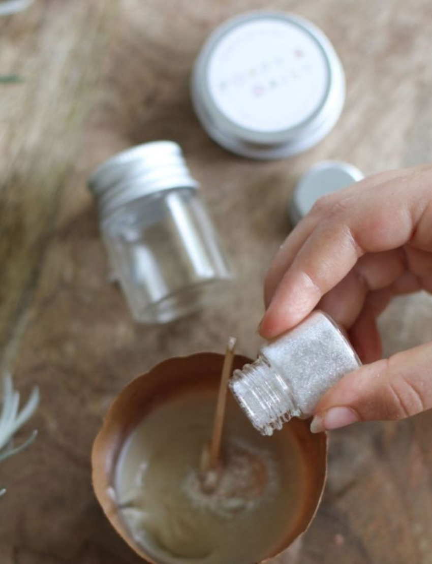 A person holds a small open tin of Natural Body Glitter while applying some to their finger. The tin is labeled "Poppy & Daisy." Pink flowers and green foliage are on the wooden table in the background.