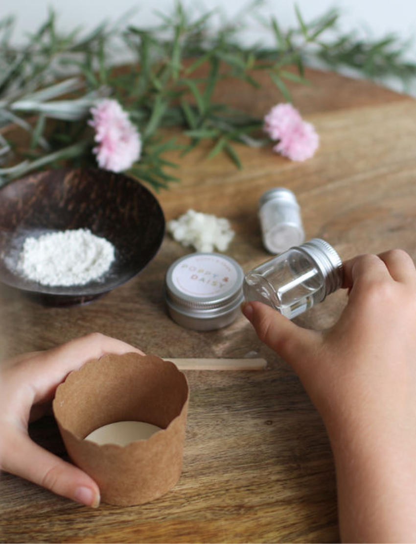 A person holds a small open tin of Natural Body Glitter while applying some to their finger. The tin is labeled "Poppy & Daisy." Pink flowers and green foliage are on the wooden table in the background.